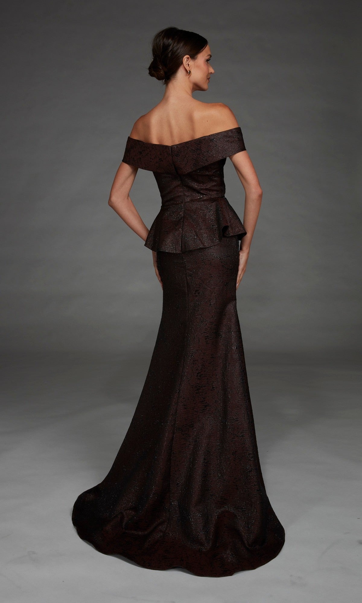 A woman models the Alyce 27892 long formal dress, featuring an off-the-shoulder neckline, peplum waist, and mermaid skirt. She stands with her back to the camera against a gray background, showcasing the gown’s elegantly draped, rich dark brown fabric that flows gracefully to the floor.